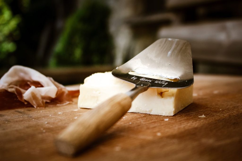 Close-up of a cheese slicer with cheese and prosciutto on a wooden board.